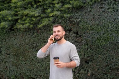 Gülümseyen adam bir telefon görüşmesi yapıyor ve bir çitin önünde dururken elinde kahve fincanı tutuyor.