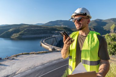Gülümseyen mühendis elinde planlar ve yenilenebilir enerji kaynağı için hidroelektrik santrali barajı yakınında akıllı telefon ses notları tutuyor.
