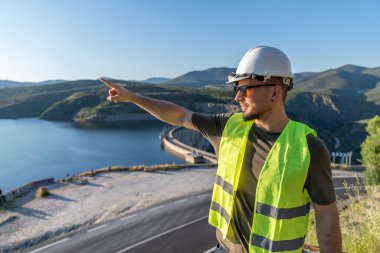 Güvenlik yeleği giyen mühendis hidroelektrik baraj ve su deposunu işaret ederek yenilenebilir enerji üretimini vurguluyor.