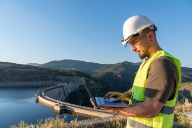 Güvenlik yeleği ve kask giyen mühendis hidroelektrik santrali barajının yanında dizüstü bilgisayar kullanarak enerji üretimini kontrol ediyor ve izliyor.