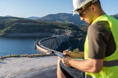 Mühendis, yenilenebilir enerji kaynağı olan hidroelektrik santrali barajının önünde notlar alıyor.