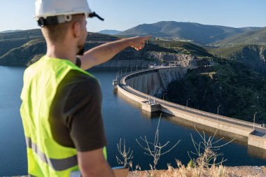 Güvenlik kaskı ve yeleği olan mühendis hidroelektrik barajını gösteriyor ve yenilenebilir enerji altyapısını gösteriyor.