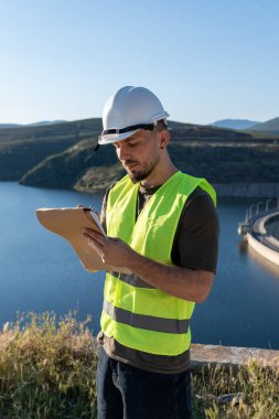 Güvenlik yeleği ve kask giyen inşaat mühendisi barajın yanındaki panoya not alıyor.