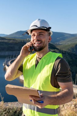 Güvenlik yeleği ve miğfer giyen mühendis telefonda konuşuyor ve dağ vadisinde bir barajın yakınındaki bir panoya not alıyor.