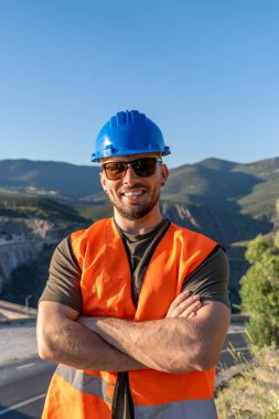 Kendine güvenen bir mühendisin portresi çapraz kollarla gülümseyen, güneş gözlüğü takan ve bir dağ yolunda şapka takan.