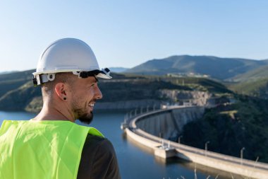 Gülümseyen mühendis çarpıcı bir dağ manzarasıyla çevrili bir baraja yansıyor, sürdürülebilir enerji ve altyapı ruhunu somutlaştırıyor.
