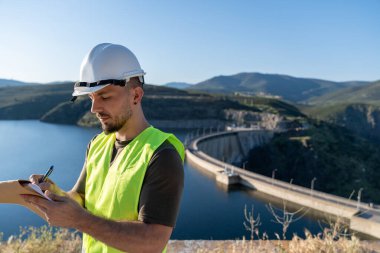 Güvenlik yeleği ve kask giyen inşaat mühendisi baraj ve su deposunun yakınındaki bir panoya not alıyor.
