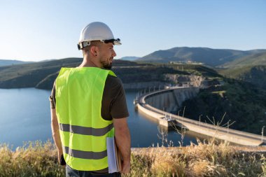 Güvenlik yeleği ve kask giyen mühendis hidroelektrik santrali barajı ve su deposunu incelerken belgeler tutuyor.