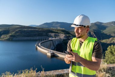 Gülümseyen mühendis hidroelektrik santrali barajının yanında cep telefonu kullanıyor, çelik yelek ve kask giyiyor, yenilenebilir enerji ve sürdürülebilirlik kavramı