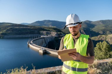 Güvenlik yeleği ve kask giyen mühendis hidroelektrik santrali barajının yakınındaki bir panoya not alıyor.