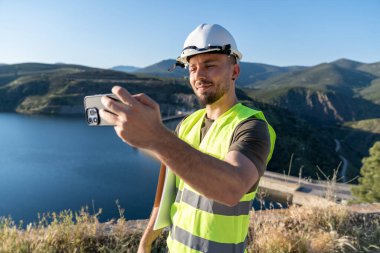 Güvenlik kaskı ve yansıtıcı yelek takmış gülümseyen mühendis akıllı telefonuyla bir su deposunun fotoğraflarını çekiyor.