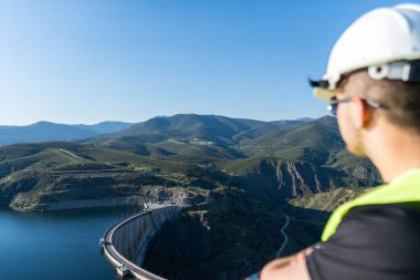 Güvenlik kaskı ve yeleği olan mühendis dağ vadisinde bir baraj ve su deposuna nezaret ediyor.