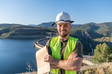 Güvenlik yeleği giyen ve kasketli bir mühendisin elinde, gülümserken, baraj ve su deposunu incelerken planları var.