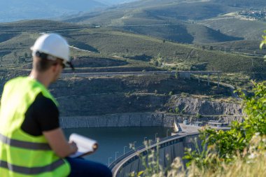 İnşaat mühendisi, barajı incelerken ve inşaat ve bakım planlama için çevredeki araziyi incelerken not alıyor.