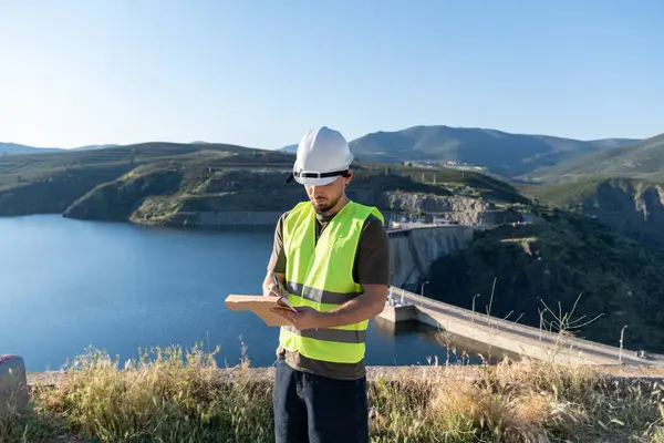 Güvenlik yeleği ve kask giyen erkek mühendis baraj ve baraj yakınındaki bir panoya not alıyor.