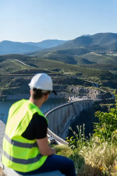 Güvenlik yeleği ve kask giyen mühendis, tepeden bir su deposu ve baraj gözlüyor, altyapı ve doğal manzaralar sergileniyor.