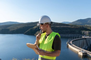 Güvenlik yeleği ve kask giyen kalite kontrolörü, bir su deposu ve hidroelektrik barajını incelerken not alıyor.
