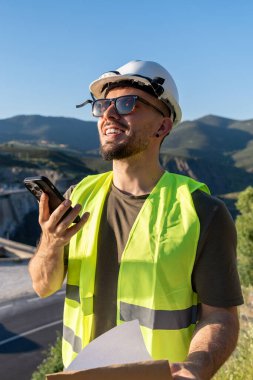 Gülümseyen kalite denetleyici güneş gözlüğü ve şapka takıyor, akıllı telefon kullanıyor ve baraj yakınlarında belgeleri tutuyor.