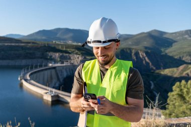 Güvenlik yeleği ve kask takan gülümseyen mühendis akıllı telefon kullanıyor ve baraj ve su deposunun yanında notlar alıyor.