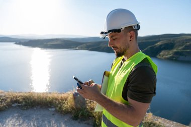 Güvenlik kaskı ve yeleği takan kalite denetleyici, güneş batarken su deposunun yakınındaki akıllı telefonla ölçümleri kontrol ediyor.