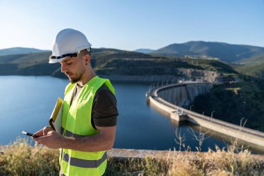 İnşaat mühendisi çelik yelek ve kask giyiyor su barajının yanında akıllı telefon kullanıyor, teftiş yapıyor ve kalite kontrolü yapıyor.