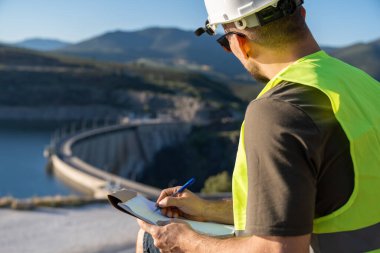 Güvenlik kaskı ve yelek giyen kalite kontrolörü barajı inceliyor ve not alıyor.