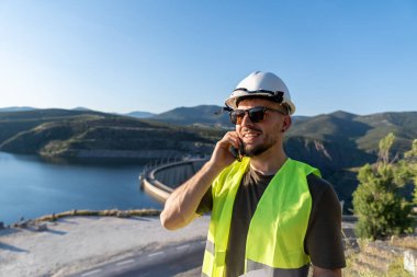Güneş gözlüğü ve şapka takan, baraj ve göl kenarında akıllı telefon kullanan, gülümseyen kalite denetleyici.