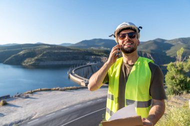 Güvenlik yeleği ve miğfer giyen mutlu mühendis, telefonla konuşuyor ve baraj inşaatını incelerken elinde planlar tutuyor.