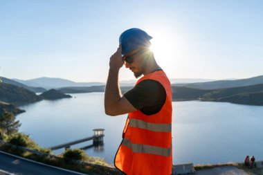 İnşaat mühendisi baraj inşaatında çalışırken sabit kaskı ayarlıyor, işyeri güvenliğini sağlıyor ve proje ilerlemesini sağlıyor