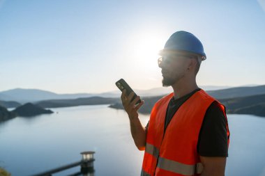 Göl kenarında akıllı telefon kullanarak güvenlik yeleği ve kask takan mühendis, sanayi ortamında iş güvenliği ve iletişim sağlıyor