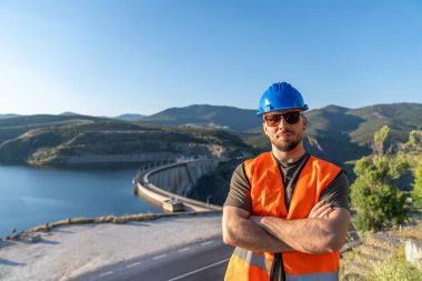 Güvenlik yeleği giyen ve kasketli kendinden emin erkek mühendis hidroelektrik santrali barajının önünde çapraz kollarla ayakta duruyor.
