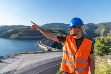 Güvenlik yeleği ve baret takan mühendis, arka planda göl olan bir dağ yolundan barajı işaret ediyor.