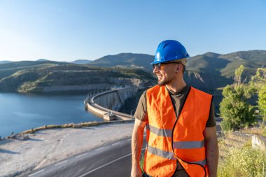 Bir hidroelektrik santrali barajını denetleyen turuncu, yüksek görüşlü yelek ve mavi başlık giyen gülümseyen bir mühendis.