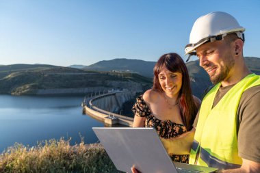 İki mühendis, doğayla çevrili bir hidroelektrik santrali barajının yanında çalışırken bir laptopta veri analizi yapıyor.