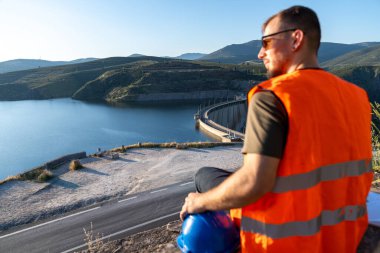 Yüksek görüş açısına sahip yelek giyen mühendis, baret tutuyor, hidroelektrik santrali ve su deposunu denetliyor, işyeri güvenliğini sağlıyor.