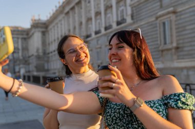 İki kadın turist, yaz tatili sırasında Madrid Kraliyet Sarayı 'nın önünde ellerinde kahve fincanları tutarken akıllı telefonlarıyla selfie çekiyorlar.