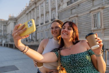 İki mutlu kadın, Madrid, İspanya 'daki kraliyet sarayının önünde akıllı bir telefonla selfie çekerken yaz tatilinin tadını çıkarıyor ve ellerinde kahve ile gezintiye çıkıyorlar.