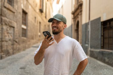 Genç adam, eski bir Avrupa şehrinde dar bir sokakta yürürken akıllı telefon ses asistanı kullanıyor.
