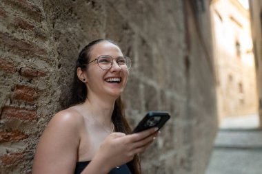 Gözlüklü genç bir kadın cep telefonu kullanıyor ve eski bir Avrupa şehrinin dar sokaklarında gülüyor.
