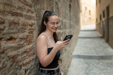 Genç bir kadın, Avrupa 'nın eski bir şehrinin dar bir sokağında duvara yaslanıp tatilinin tadını çıkarırken akıllı telefon kullanıyor.