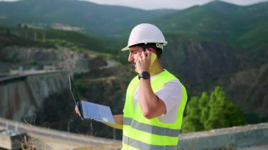 Emniyet yeleği giyen mühendis dizüstü bilgisayar kullanıyor ve telefonda ağır çekimde konuşuyor.