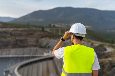Güvenlik yeleği ve başlık takan mühendis, dağlık bir bölgede verimli su kaynakları yönetimi ve yenilenebilir enerji üretimi sağlayan bir barajı denetliyor.