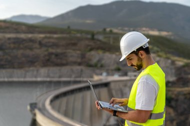 Güvenlik yeleği ve kask giyen mühendis hidroelektrik barajında çalışırken dizüstü bilgisayar kullanıyor. Su kaynaklarını ve yenilenebilir enerji kaynaklarını denetliyor.