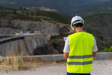Güvenlik yeleği ve kask giyen mühendis, dağlık bir bölgedeki barajı incelerken bir panoya not alıyor.