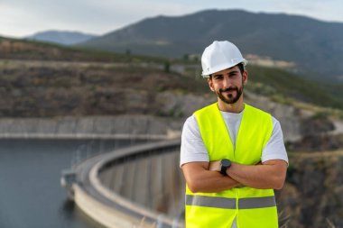 Çapraz kollu çelik yelek ve kask giyen mühendis, dağlık bir bölgedeki baraj ve su deposunu gözetliyor.