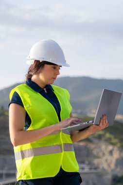 Güvenlik yeleği ve kask giyen genç kadın mühendis dizüstü bilgisayarla altyapı projesini yönetiyor.