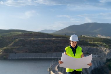 Güvenlik yeleği ve kask giyen kadın mühendis baraj barajının yakınındaki planı inceliyor, altyapı ve su yönetimi üzerinde çalışıyor.