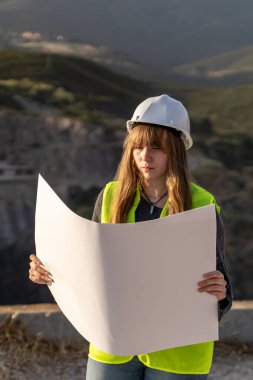 Güvenlik kaskı ve yeleği takan genç kadın mühendis, bir taş ocağının planlarını dikkatle inceliyor, proje uyumunu sağlıyor.