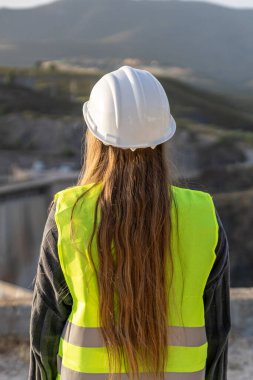 Uzun saçlı kadın mühendis, güvenlik kaskı ve yansıtıcı yelek giyiyor, bir inşaat alanını teftiş ediyor.