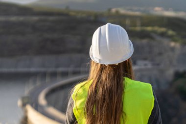 Güvenlik kaskı ve yelek giyen kadın mühendis bir barajı teftiş ederek iş yerinde iş güvenliği ve risk önlemeyi garanti altına alıyor